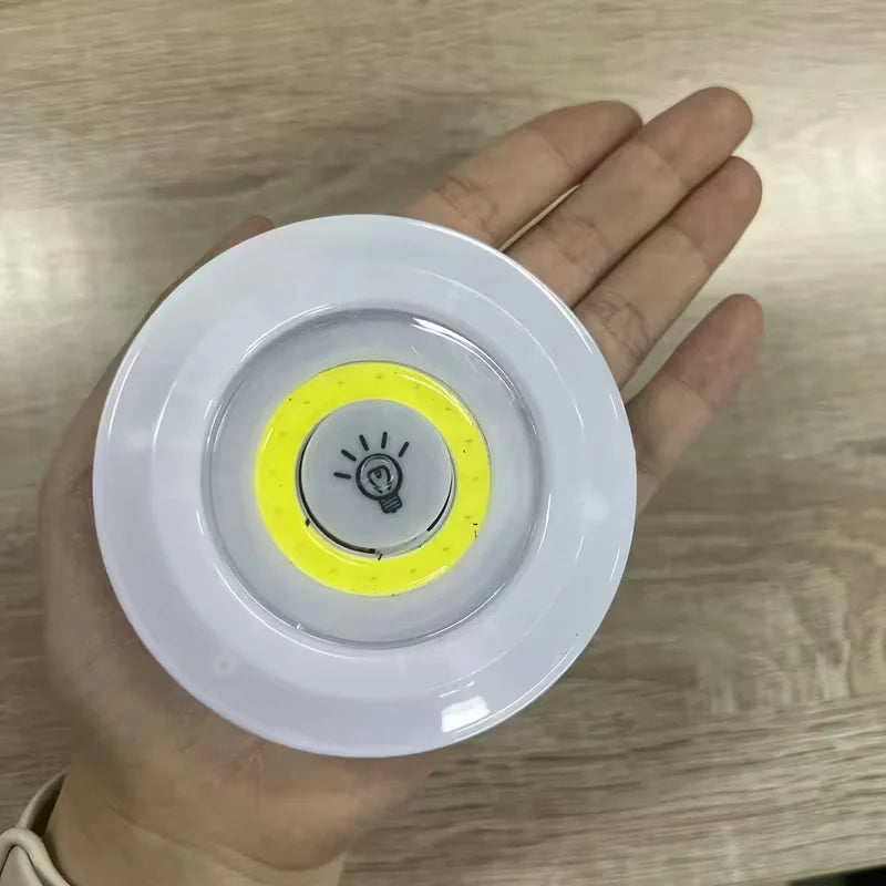 A hand holding a small round white device with a yellow ring and a light bulb symbol in the center, photographed over a wood-grain surface.