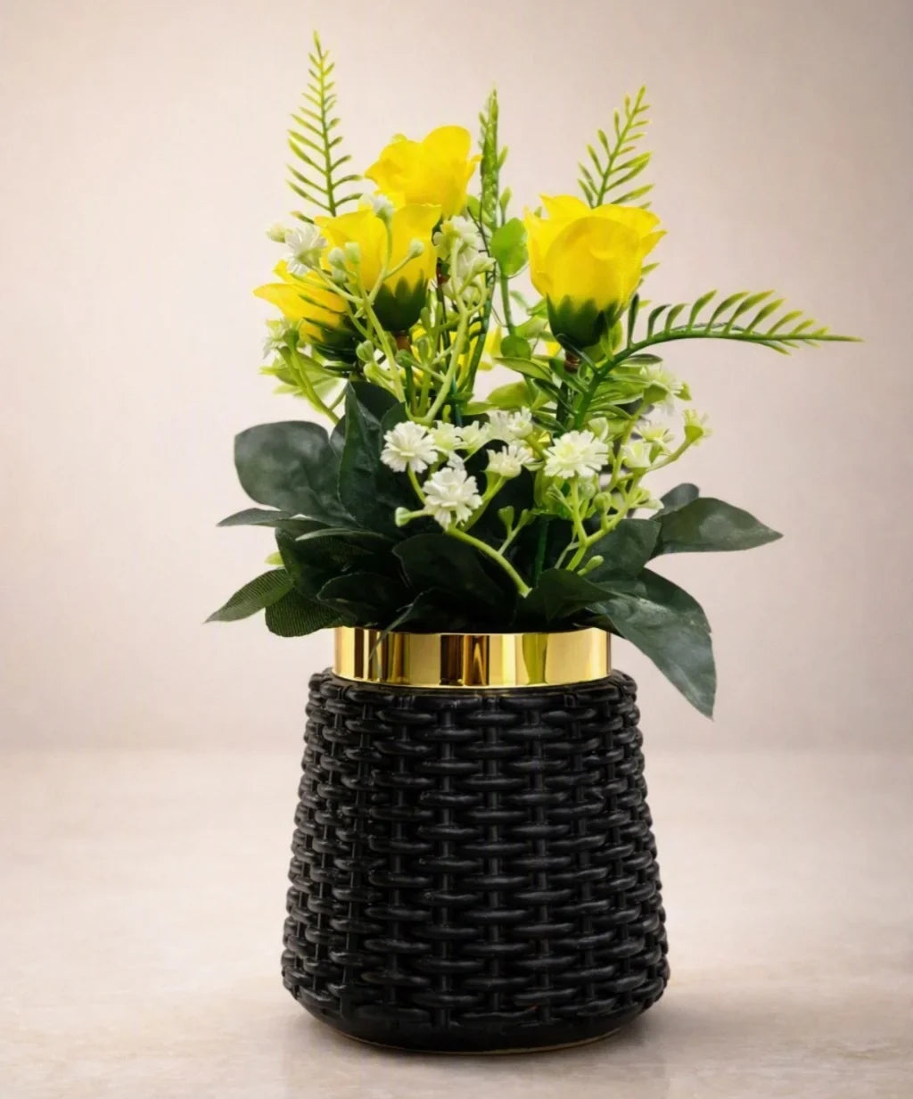 A decorative artificial flower arrangement with bright yellow roses, green foliage, and small white blossoms in a black woven vase with a gold band, set against a plain beige background.