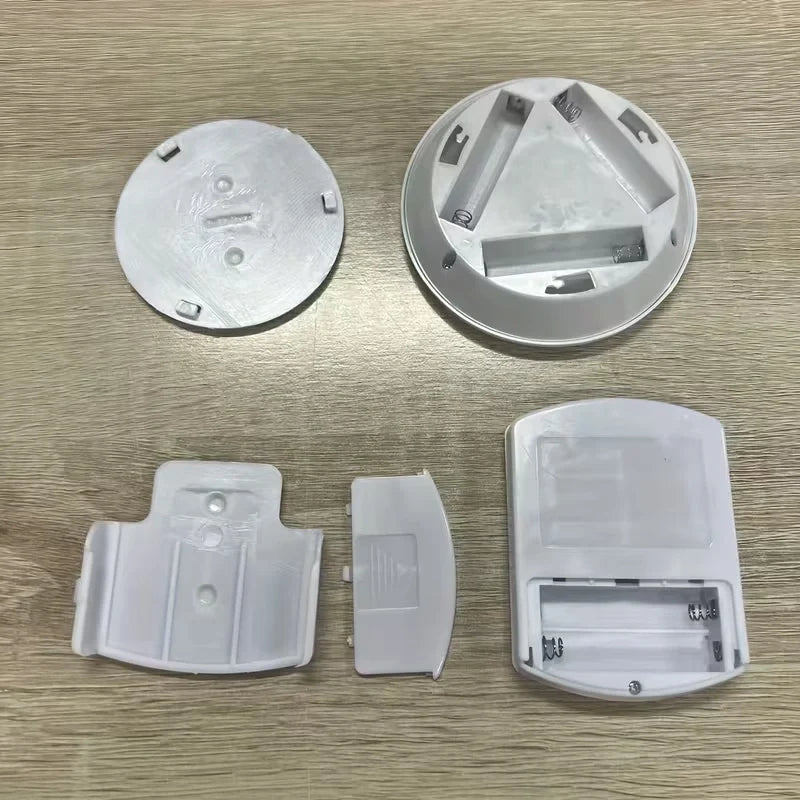 A set of white plastic replacement parts for a circular electronic device, laid out on a light wood-grained surface. The parts include a circular base, a circular top with battery compartments, a curved cover, and a small rectangular piece.