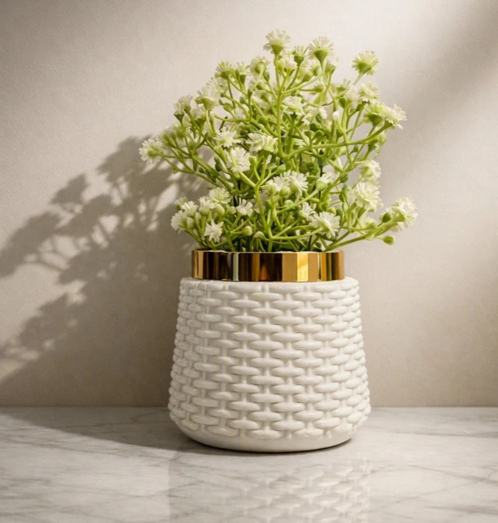 A white textured ceramic planter with a gold band around the rim, holding white baby's breath flowers, set on a light marble surface against a beige wall with soft shadows.