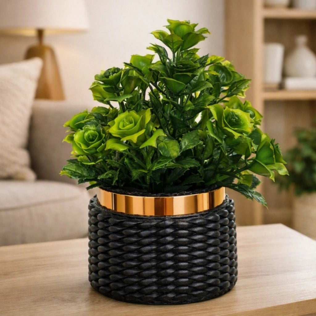 An artificial rose bush with vibrant green faux roses and leaves, presented in a black woven planter with a metallic gold band, sitting on a wooden table in a cozy living room setting.