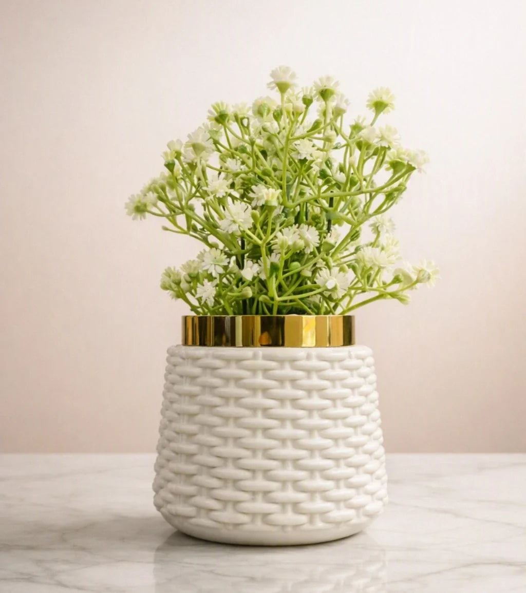 A white textured ceramic planter with a gold metallic rim, filled with small white flowers and green stems, set on a light marble surface against a soft beige background.