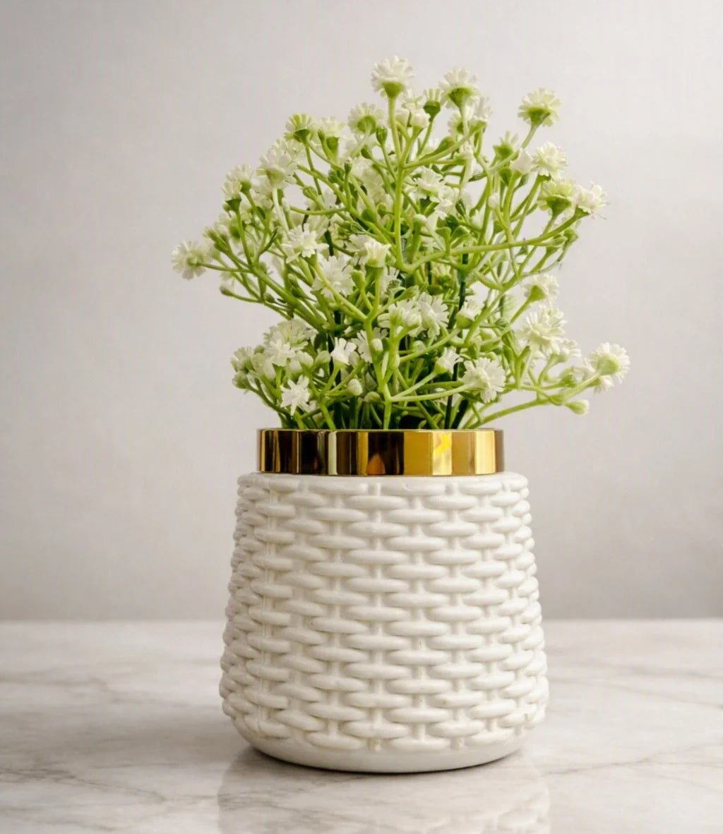 A white ceramic planter with a textured woven basket pattern and a shiny gold rim, filled with small white flowers and green stems, set on a light marble surface against a neutral gray background.