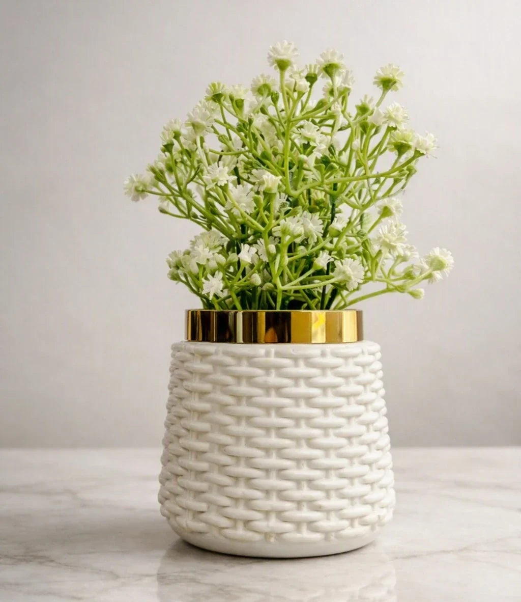 A white ceramic planter with a textured woven basket pattern and a shiny gold rim, filled with small white flowers and green stems, set on a light marble surface against a neutral gray background.