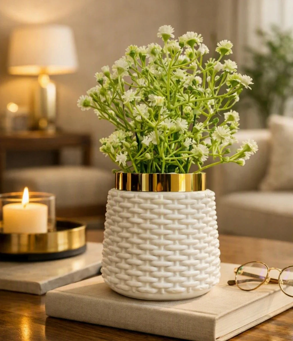 A white textured ceramic planter with a gold rim, filled with small white flowers, styled on a book on a wooden table in a warm, softly lit living room.