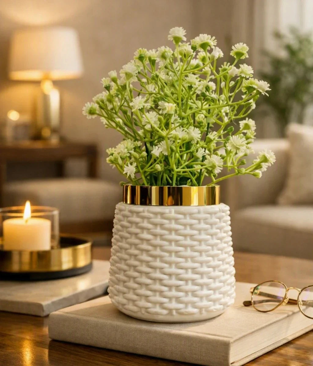 A white textured ceramic planter with a gold rim, filled with small white flowers, styled on a book on a wooden table in a warm, softly lit living room.