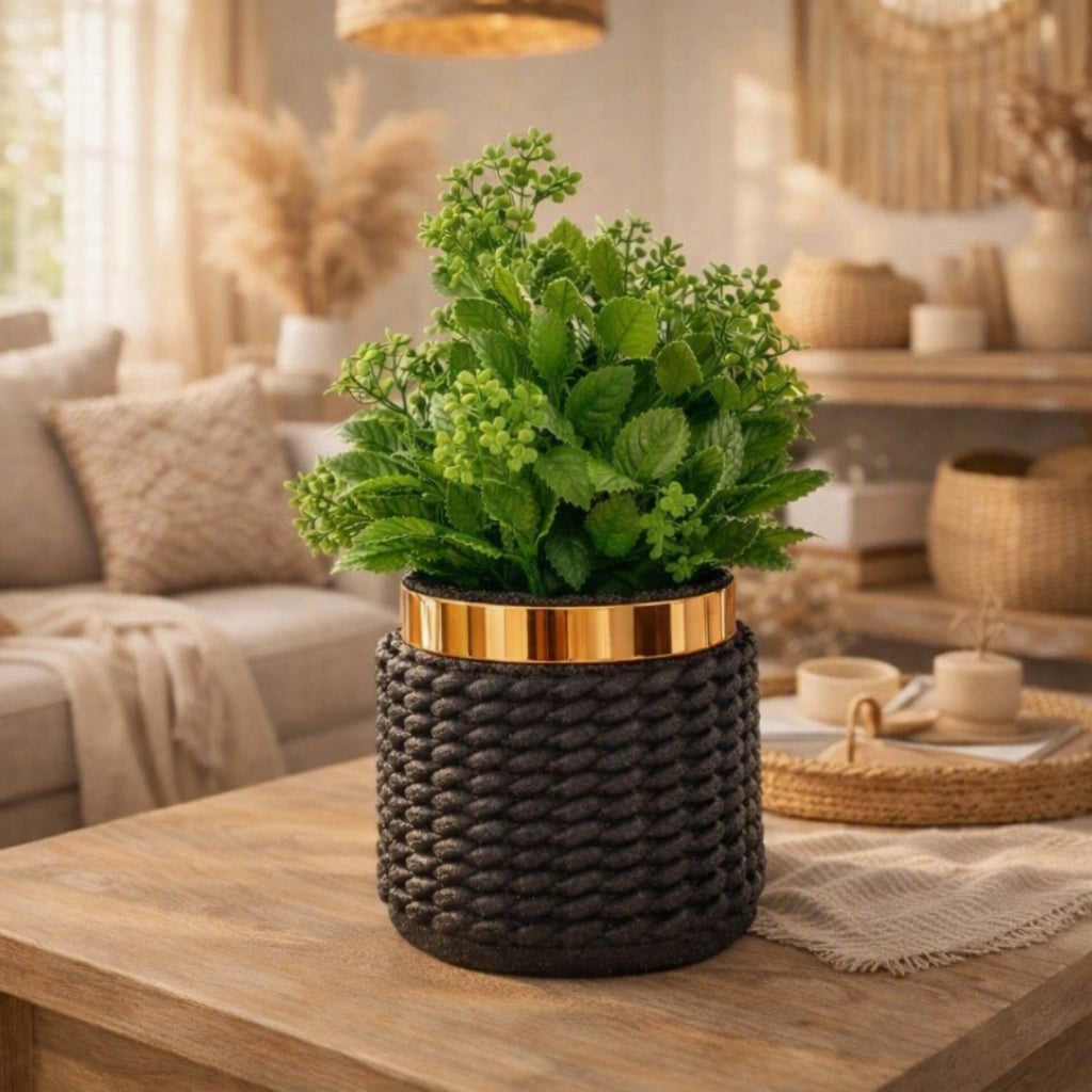An artificial green leafy plant in a dark woven cylindrical pot with a metallic gold band, displayed on a wooden table in a warm, modern living room setting.