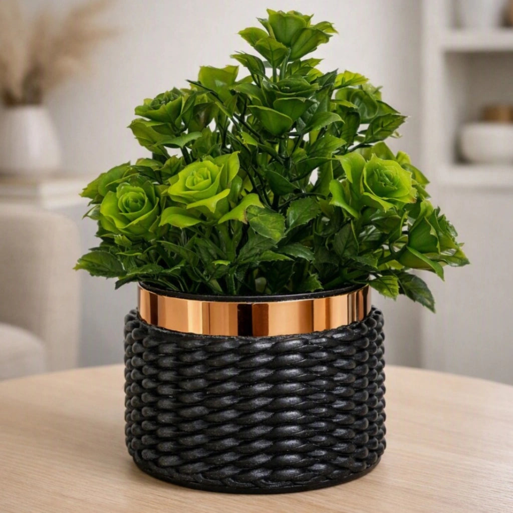 A potted artificial rose plant with lush green faux blooms in a black woven basket-style pot with a metallic copper-gold rim, set on a light wood table in a softly blurred indoor setting.