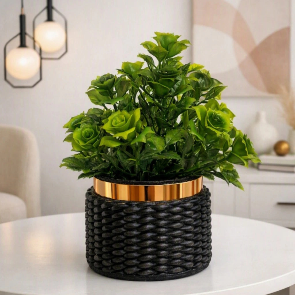 A green artificial rose plant in a black woven planter with a metallic gold band, displayed on a white table in a modern living room setting.