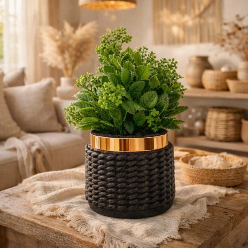 A potted artificial green plant with dense leafy stems sits in a dark woven cylindrical planter with a metallic gold band, placed on a wooden table in a softly lit living room setting.