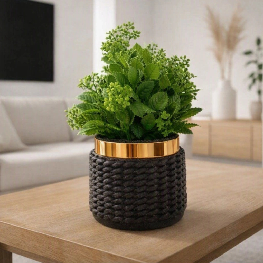 A green leafy plant in a dark woven basket-style pot with a metallic gold band, placed on a light wood table in a softly blurred living room setting.