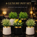 Six luxury decorative potted plants arranged on a dark marble surface in a warmly lit interior setting. The set includes three woven white pots with yellow flowers, two black woven pots with green foliage, and two black pots with white flowers and greenery.