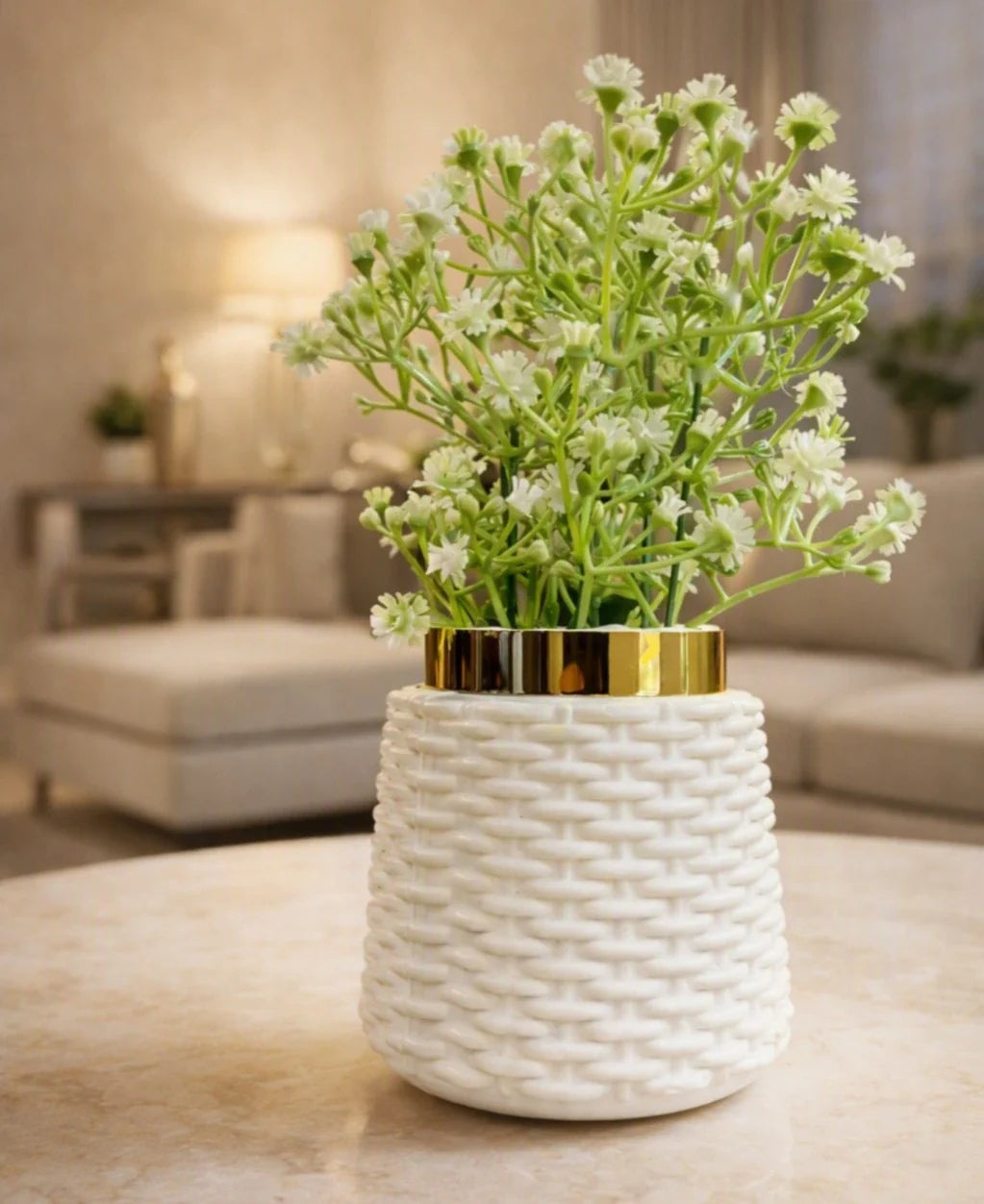 A white woven-texture ceramic vase with a shiny gold rim, filled with small white flowers, set on a light marble table in a softly blurred living room background.