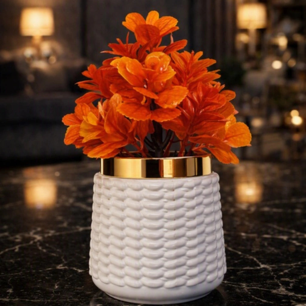 A decorative artificial orange flower arrangement in a textured white cylindrical pot with a metallic gold band, placed on a dark marble surface in a softly blurred indoor setting.