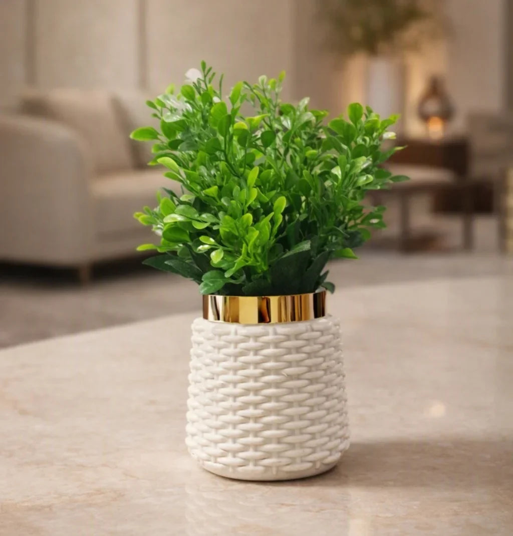 A small potted artificial green plant with dense leafy foliage, set in a white woven ceramic pot with a shiny gold band at the rim, placed on a light marble tabletop in a softly blurred living room background.