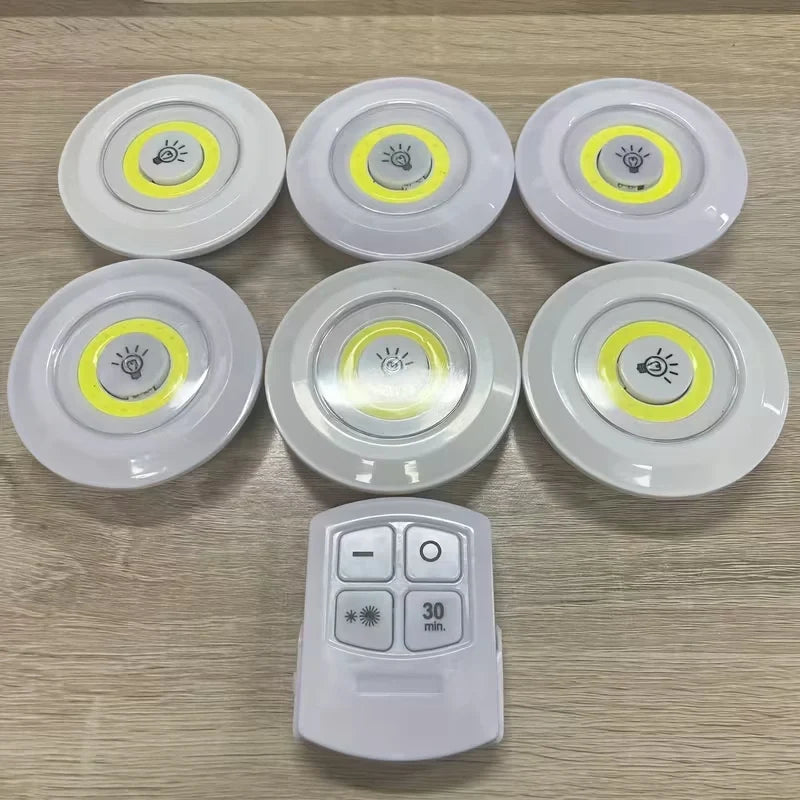 Six round white LED under-cabinet light sensors with yellow accent rings are arranged on a light wood surface, with a white remote control placed in front.